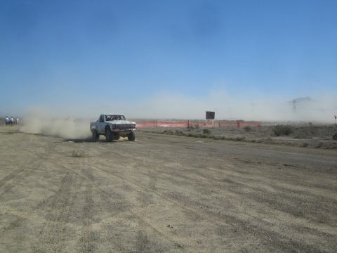1990 Ford Bronco - white off-road race truck speeds across an open dirt course, leaving a trailing dust cloud under a clear blue sky.