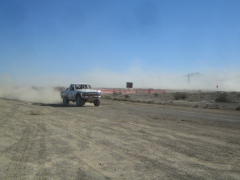 1990 Ford Bronco - white off-road race truck rounds a bend on a dusty desert dirt course beside a red barrier fence.