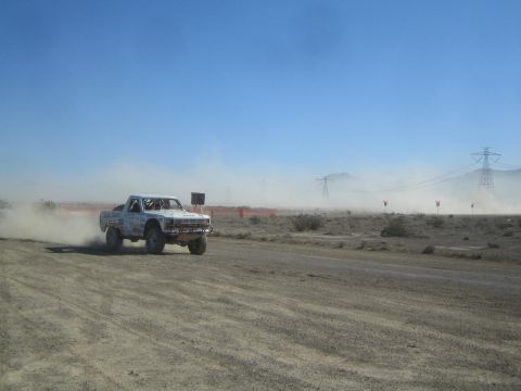 1990 Ford Bronco - white off-road race truck races along a dirt road with transmission towers and dust in the distance.