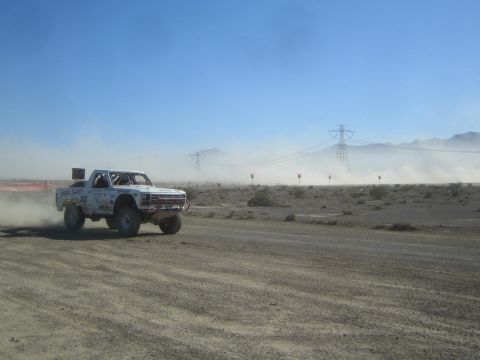 1990 Ford Bronco - white off-road race truck catches air over a rise on a desert dirt course with mountains and power lines behind.