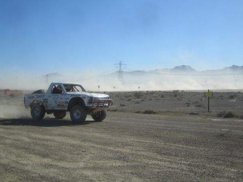 1990 Ford Bronco - white off-road race truck marked 302 speeds along a dirt course in the desert with mountains in the background.