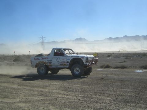 1990 Ford Bronco - white off-road race truck lettered Anger Issues and numbered 302 races across a dusty desert course toward distant mountains.