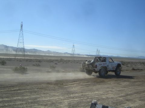 1990 Ford Bronco - white off-road race truck races across open desert terrain under tall transmission towers and a blue sky.
