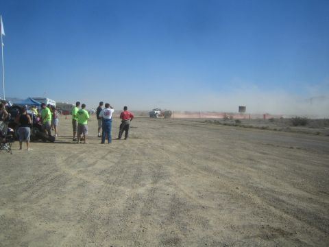 1990 Ford Bronco - group of spectators stands at a dusty desert course watching a race truck kick up a dust cloud in the distance.