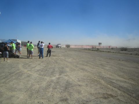 1990 Ford Bronco - Spectators gather near tents along a dusty desert dirt course as a race truck raises dust far down the track.