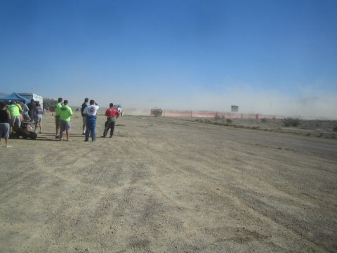 1990 Ford Bronco - Spectators stand at the edge of a desert dirt course watching a race vehicle raise a large dust cloud in the distance.