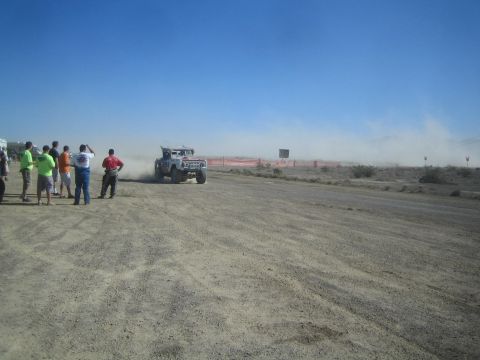 1990 Ford Bronco - white off-road race truck kicks up dust as it accelerates past a line of spectators on a desert dirt course.
