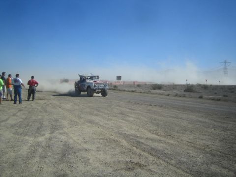 1990 Ford Bronco - white tube-frame off-road race truck races past spectators on a desert dirt course with power lines behind.