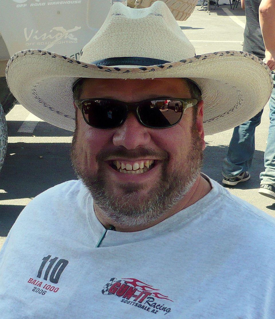 Man in cowboy hat and sunglasses smiles, wearing a shirt with 'Baja 1000 2006' and 'Gon-Fit Racing' logos