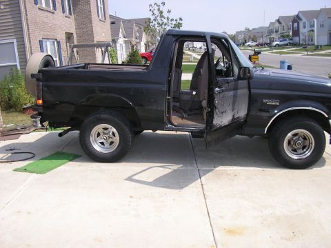 1996 Ford Bronco - Passenger-side profile of a black full-size Ford Bronco in a suburban driveway with the rear hardtop removed to leave an open bed, the pass…