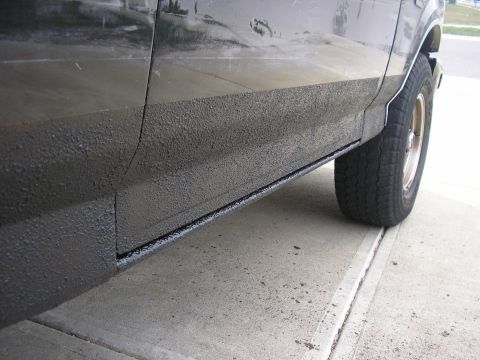 1996 Ford Bronco - Low-angle close-up of the lower body side of a dark truck on a concrete driveway, showing a freshly applied textured black bedliner-style c…