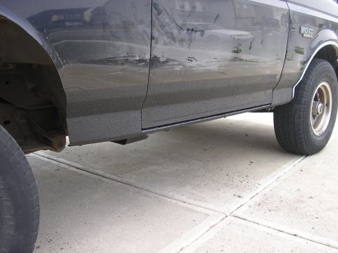 1996 Ford Bronco - Passenger-side low view of a dark Ford Bronco on a concrete driveway with textured black bedliner coating rolled along the rocker panel, lo…