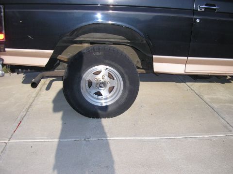 1996 Ford Bronco - The rear wheel and lower body panel of a dark SUV are visible, showing tan trim and a silver rim.