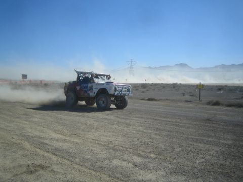 1990 Ford Bronco - white-and-blue open-bodied off-road race truck rounds a bend on a dusty desert course beneath a transmission tower.
