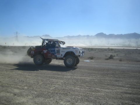1990 Ford Bronco - white and red off-road race truck numbered 305 races across a desert dirt course with mountains in the distance.
