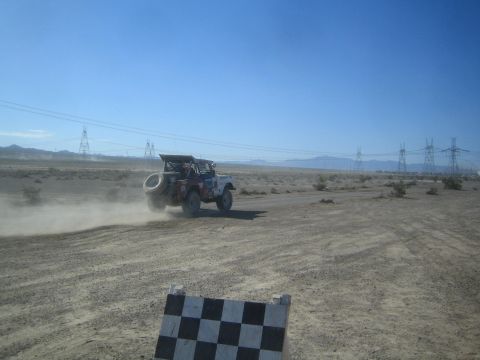 1990 Ford Bronco - off-road race truck with a rear-mounted spare races across the desert past a black-and-white checkered course marker.