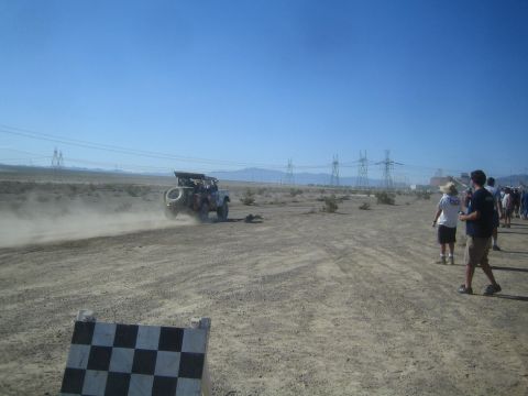 1990 Ford Bronco - Spectators stand beside a desert course watching an off-road race truck raise dust near a checkered marker board.