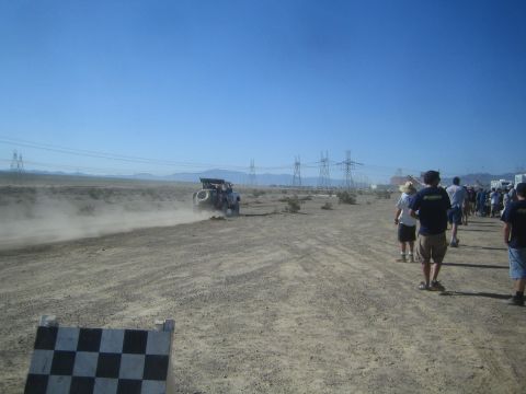 1990 Ford Bronco - Spectators walk along a dusty desert dirt course while an off-road race truck speeds by near a checkered marker.