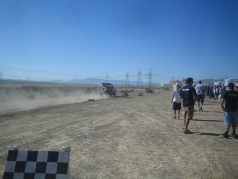 1990 Ford Bronco - Spectators walk a desert dirt course as a distant off-road race truck trails dust under transmission towers.