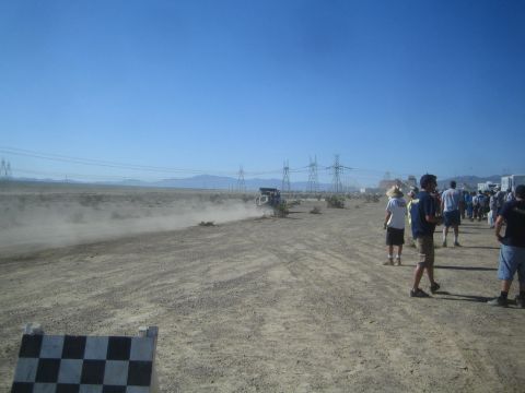 1990 Ford Bronco - Crowd of spectators lines a dusty desert dirt track as a distant off-road vehicle kicks up a dust trail under power lines.
