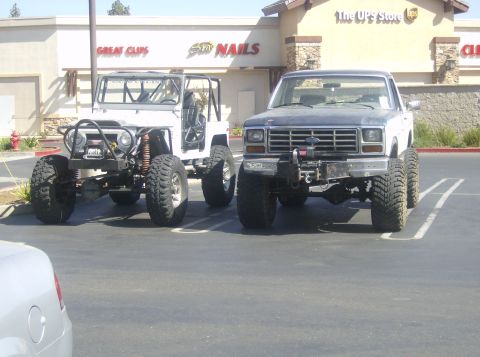 1986 Ford Bronco - Two heavily modified trucks are parked in a lot, one a Ford pickup and the other a white off-road vehicle.