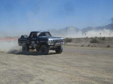 1990 Ford Bronco - Black West Coast Broncos off-road race truck number 300 throws up dust while racing along a desert course past red marker flags.