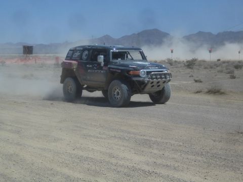 1990 Ford Bronco - Black Toyota FJ Cruiser built for off-road racing kicks up dust on a desert course backed by mountains.