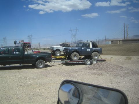 1990 Ford Bronco - Green pickup numbered 348 tows a trailer carrying two early Ford Bronco race rigs in a gravel desert lot.