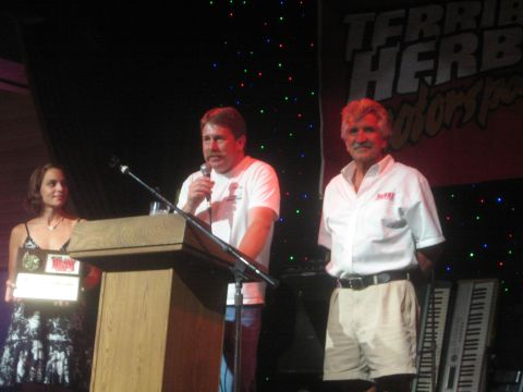 1990 Ford Bronco - Man speaks into a microphone at a podium between two people on a darkened stage under a Terrible Herbst Motorsports banner.