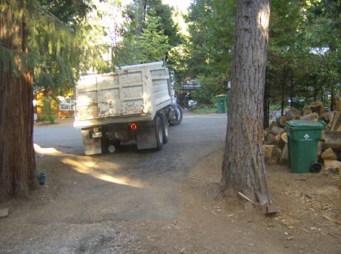 1986 Ford Bronco - A dump truck is parked on a dirt and asphalt surface near trees and houses.