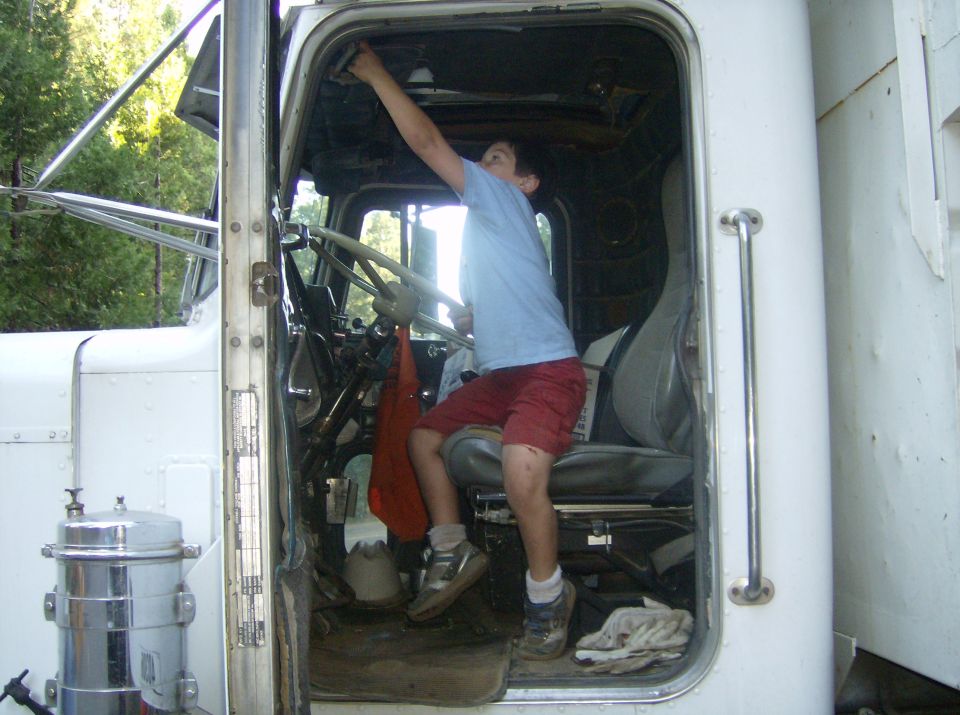 Boy in red shorts and blue shirt reaching for the ceiling inside a semi-truck cab