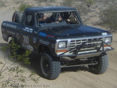 1990 Ford Bronco - Blue West Coast Broncos race truck number 300 with two occupants drives across desert sand with a tube front bumper.
