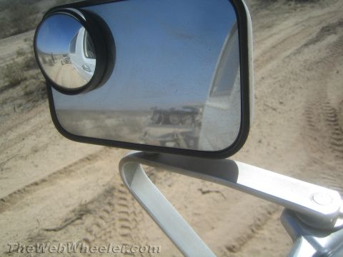 1990 Ford Bronco - Round convex spot mirror mounted on a tubular arm reflects a dusty desert trail and trailing vehicle.