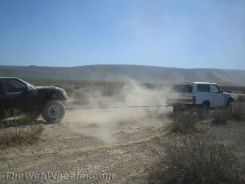 1990 Ford Bronco - Dark off-road truck tows a white full-size Ford Bronco via a strap, raising dust on a desert trail.