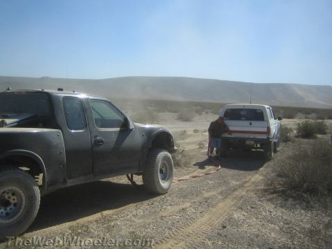 1990 Ford Bronco - Dusty black pickup is connected by a tow strap to a white Ford rig while a person crouches to attach it in the desert.