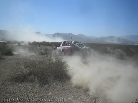 1990 Ford Bronco - Red and white off-road race truck blasts through desert brush, throwing up a large cloud of dust against distant mountains.