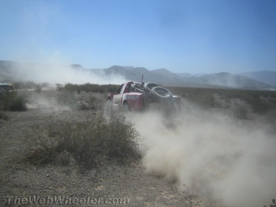 Red and white off-road race truck powering through desert brush in dust