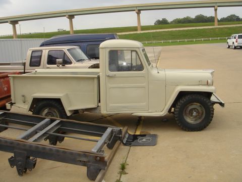 1958 Jeep J-10 - A cream-colored pickup truck is parked outdoors, with other vehicles and a highway overpass in the background.