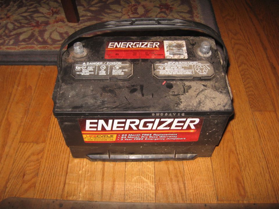 Dirty black Energizer Group 65 car battery with carry strap sitting on a hardwood floor
