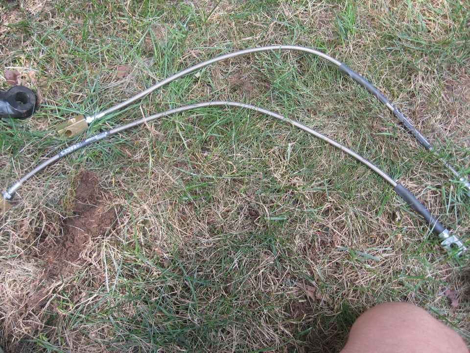 Two stainless steel brake lines with black protective sleeves and brass fittings lie on grass