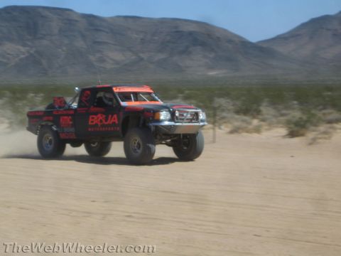 1990 Ford Bronco - Black and orange Boja Motorsports off-road race truck speeds across an open desert flat, trailing dust.