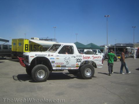 1990 Ford Bronco - White off-road Bronco race truck number 302 with sponsor decals sits in a paved pit area as people walk past.