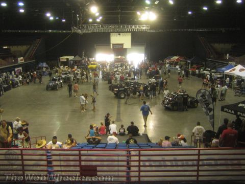1990 Ford Bronco - Spectators in arena seating overlook a show floor displaying off-road race vehicles and PIAA vendor booths under bright lights.
