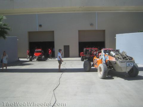 1990 Ford Bronco - Orange number 117 and red tube-chassis off-road buggies sit on a paved lot outside warehouse bay doors as crew members stand by.