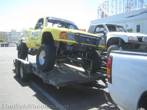 1990 Ford Bronco - Yellow lifted Ford pickup prerunner-style race truck secured on a tandem-axle flatbed trailer in a sunny lot.
