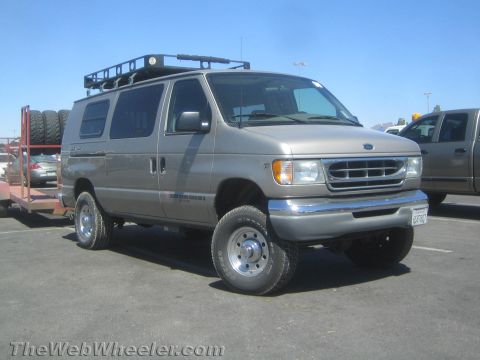 1990 Ford Bronco - Gold lifted Ford E-series van with roof rack and oversized tires parked beside a trailer in a sunny lot.