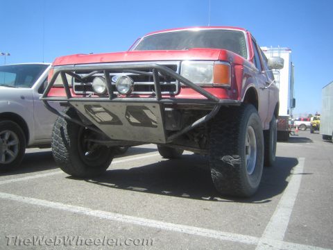 1990 Ford Bronco - Low front view of a red lifted Ford truck with a fabricated tube bumper, dual driving lights and chunky off-road tires.