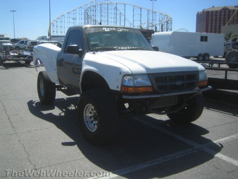 1990 Ford Bronco - Front three-quarter view of a white lifted Ford Ranger prerunner truck with fiberglass fenders, parked near a roller coaster.