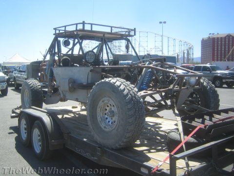 1990 Ford Bronco - Rear view of a bare tube-chassis off-road buggy with roll cage and big tires loaded on a tandem-axle trailer near a fairground.