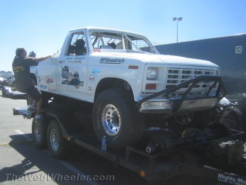 1990 Ford Bronco - White lifted Ford F-series race truck with BFGoodrich and sponsor decals on a flatbed trailer while a man climbs up beside it.